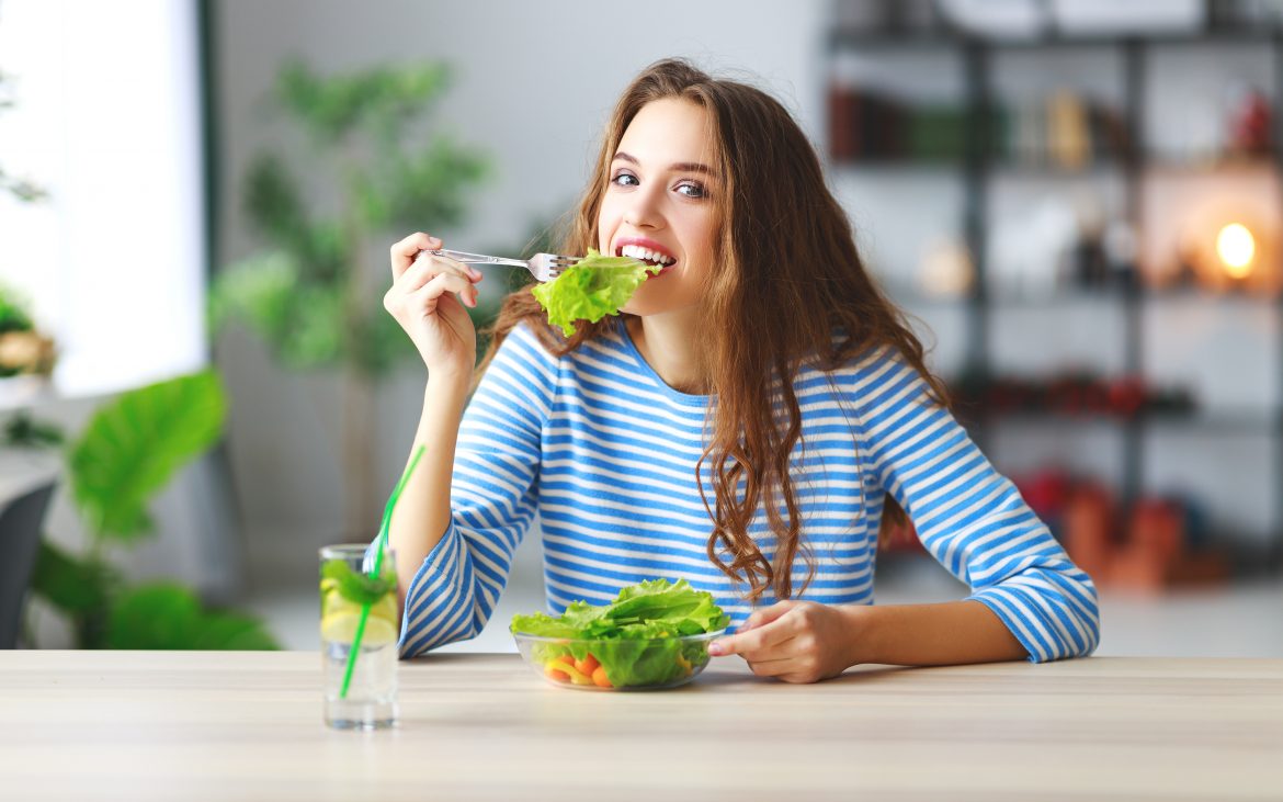 Dieta przy PCOS - co jeść, gdy cierpimy na zespół policystycznych jajników? Młoda kobieta w bluzce w biało-niebieskie paski je sałatkę, siedząc przy stole kuchennym.