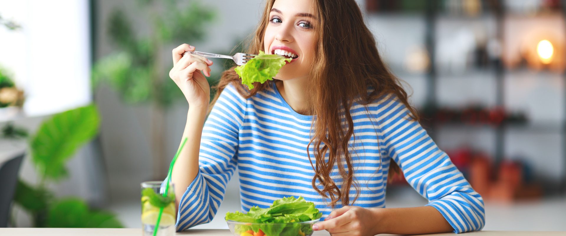 Dieta przy PCOS - co jeść, gdy cierpimy na zespół policystycznych jajników? Młoda kobieta w bluzce w biało-niebieskie paski je sałatkę, siedząc przy stole kuchennym.