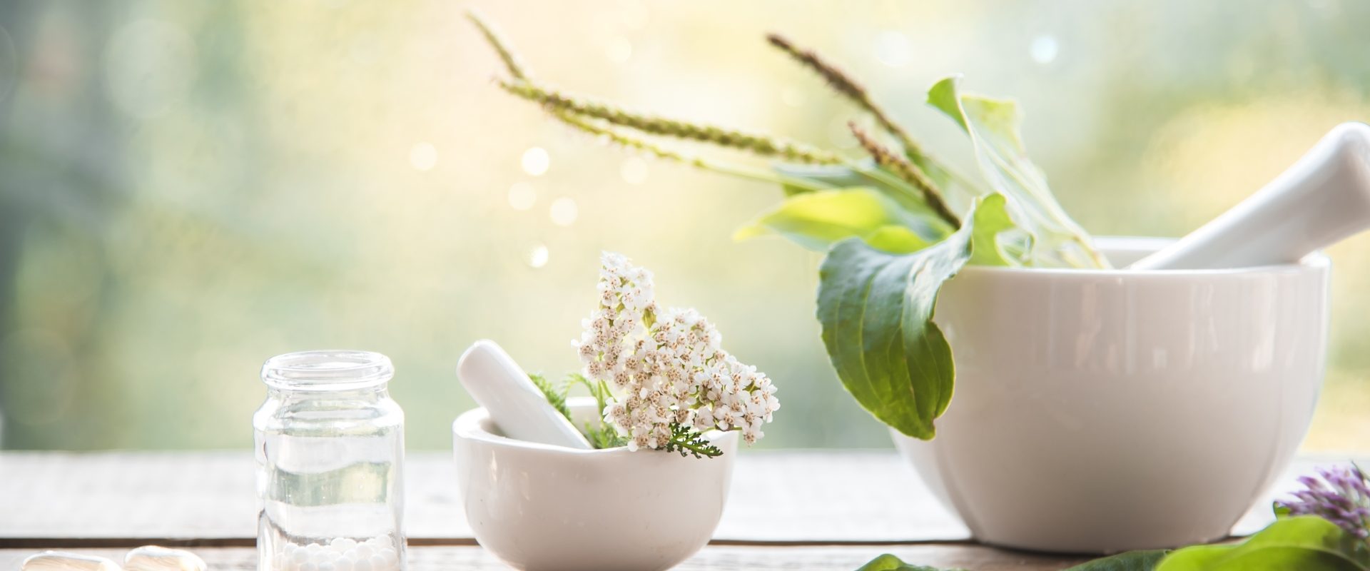 Lek homeopatyczny Phosphorus na energię i zwiększenie sił witalnych. Liście roślin w białym moździerzu stojącym na stole w ogrodzie, obok leżą granulki leków homeopatycznych.