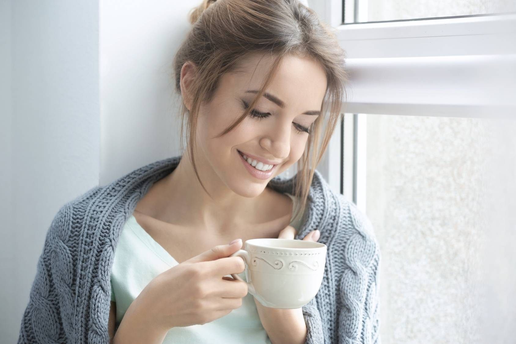 Phytolacca decandra - lek homeopatyczny na ból gardła. Kobieta otulona swetrem siedzi na parapecie i pije herbatę z białej filiżanki.