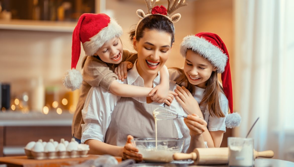 Jak zrobić pierniczki świąteczne? Mama z córkami w czapkach Świętego Mikołaja pieką pierniczki na Święta w domowej kuchni.