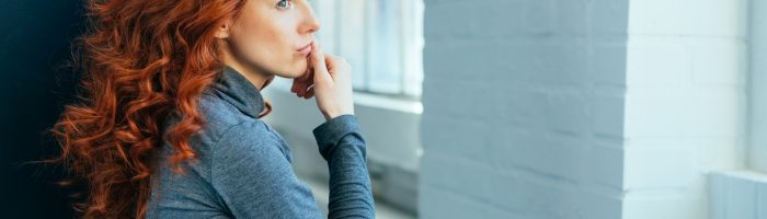 Jakie homeopatyczne leki na PMS mogą ci pomóc? Smutna kobieta w gęstych kręconych rudych włosach, w szarym golfie, zastanawia się nad czymś, patrząc w okno.