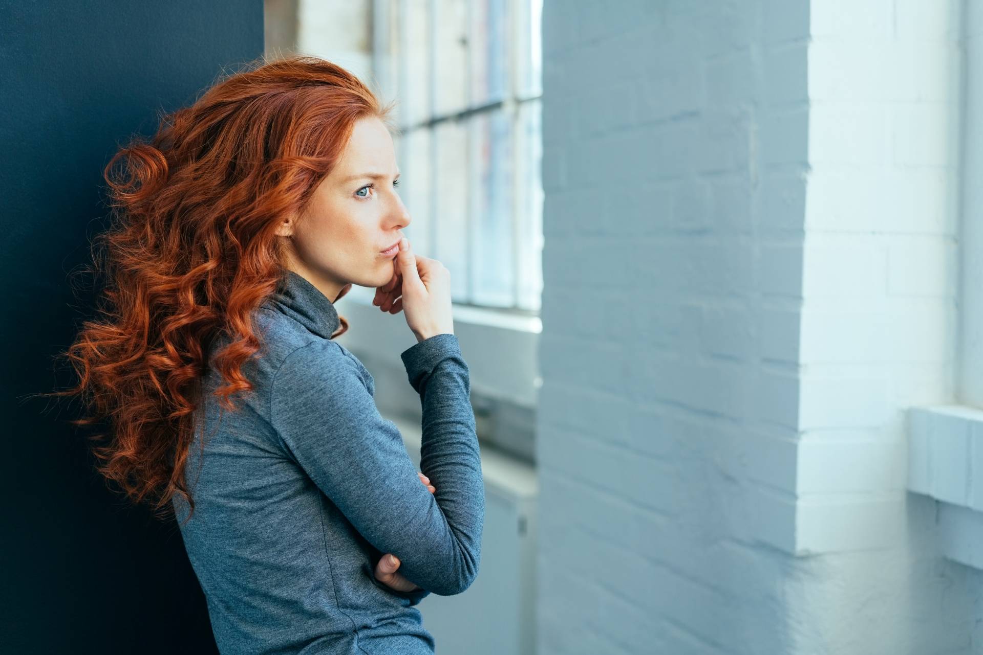 Jakie homeopatyczne leki na PMS mogą ci pomóc? Smutna kobieta w gęstych kręconych rudych włosach, w szarym golfie, zastanawia się nad czymś, patrząc w okno.