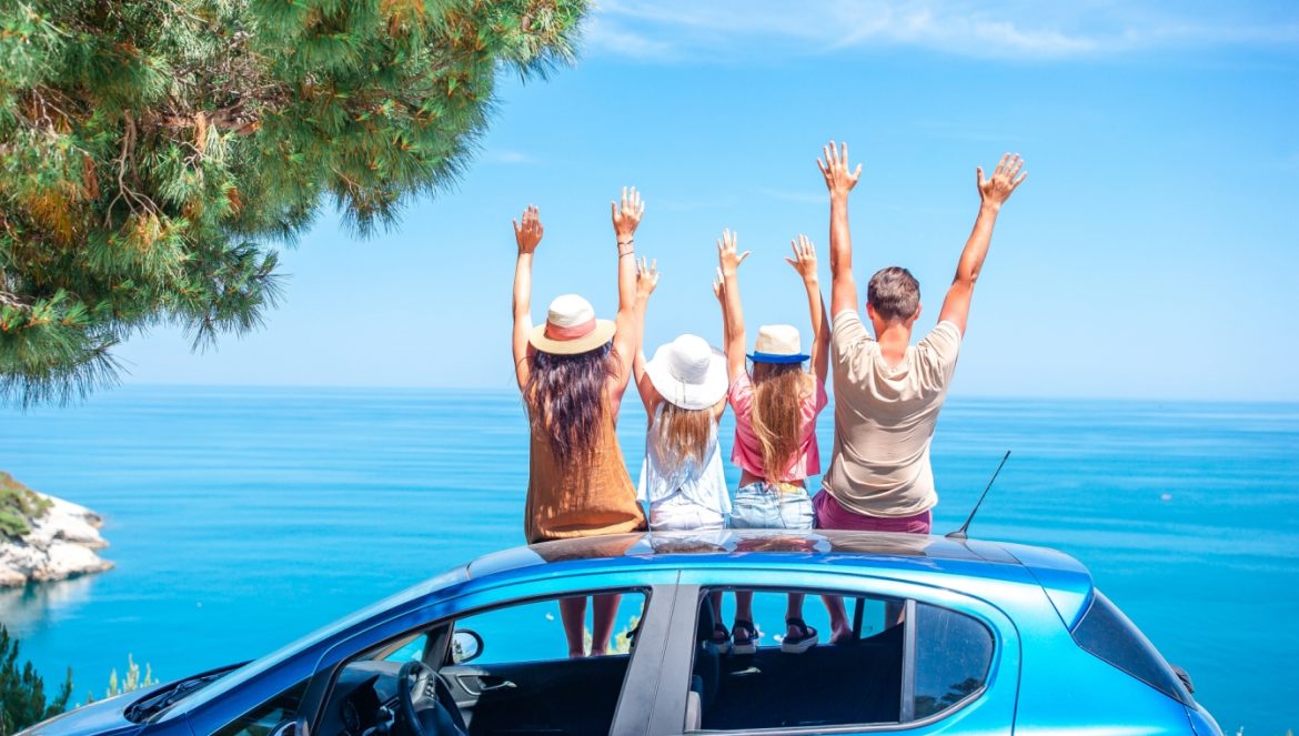 Beata Sadowska: podróże z dziećmi nie takie straszne, jakby się mogło wydawać. Dziennikarka i podróżniczka opowiada o tym, w co się bawi z dziećmi w podróży. Szczęśliwa rodzina na wakacjach siedzi na dachu samochodu z wyciągniętymi do góry rękami i podziwia widok na morze.