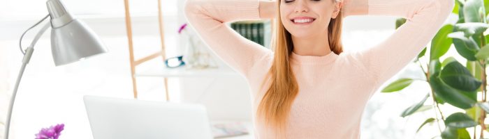 Wyluzuj, czyli jak wyzbyć się perfekcjonizmu. Jak zdystansować się do pracy i obowiązków i ciągłej potrzeby bycia najlepszym? Uśmiechnięta zrelaksowana kobieta siedzi przy biurku z rękami założonymi za głowę.