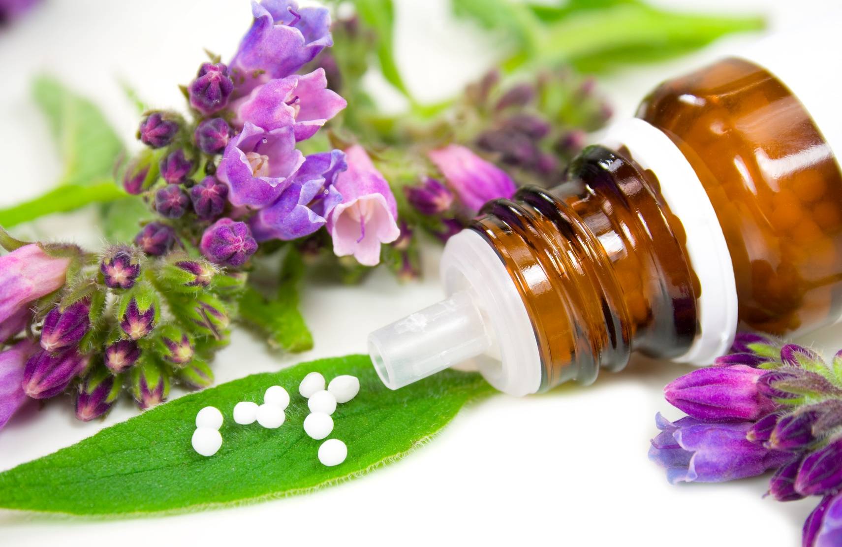 Homeopatia, lek homeopatyczny Ignatia amara - dla kogo i na co pomaga? Fiolka leku homeopatycznego w otoczeniu fioletowych kwiatów i zielonych liści.