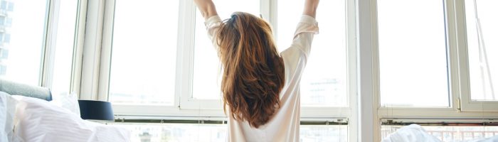 Praktyka i medytacja wdzięczności, czyli jak dobrze zacząć dzień. Młoda kobieta w białej koszuli wstaje z łóżka o poranku. siedząc jeszcze na łóżku, przeciąga się patrząc w okna.