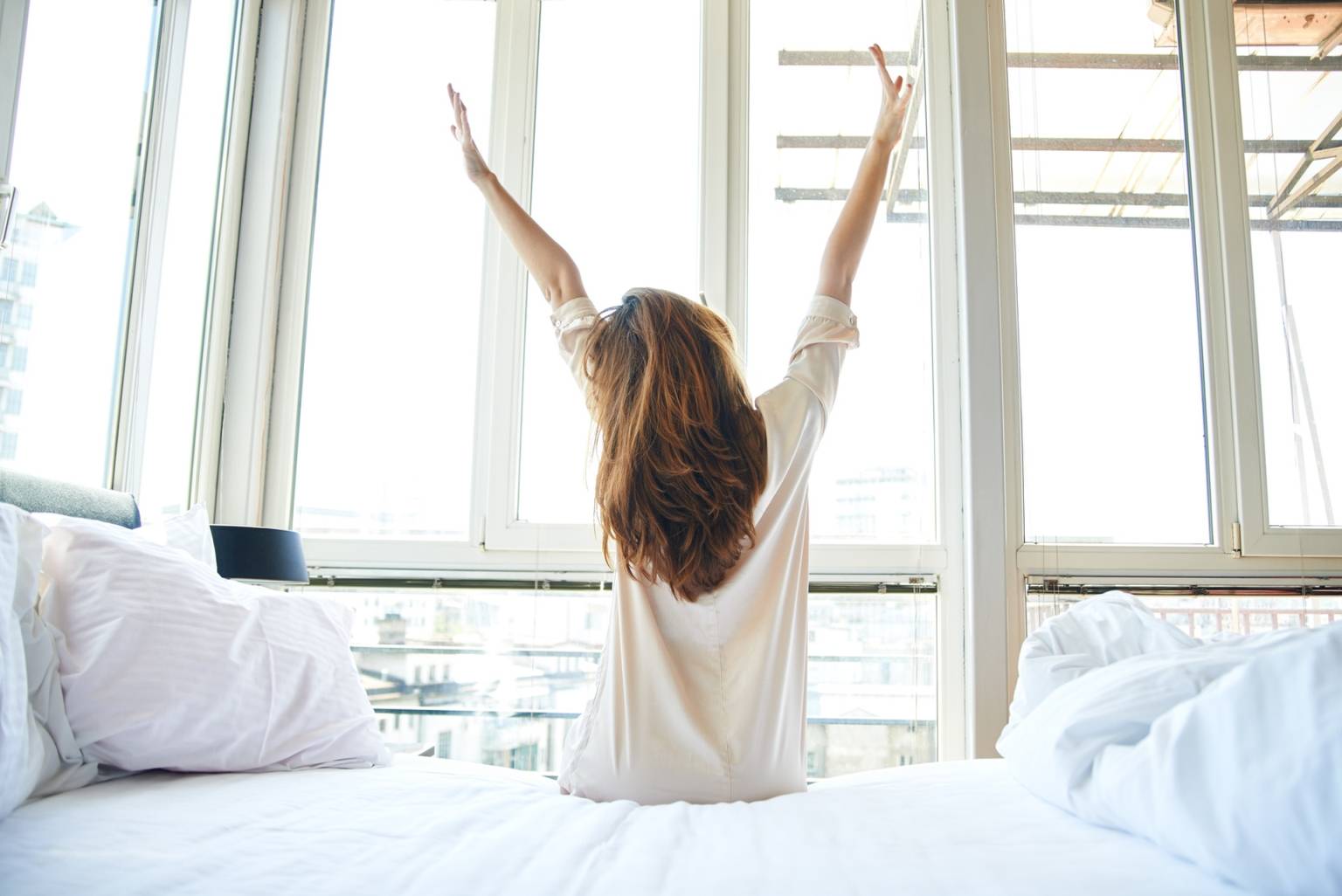 Praktyka i medytacja wdzięczności, czyli jak dobrze zacząć dzień. Młoda kobieta w białej koszuli wstaje z łóżka o poranku. siedząc jeszcze na łóżku, przeciąga się patrząc w okna.