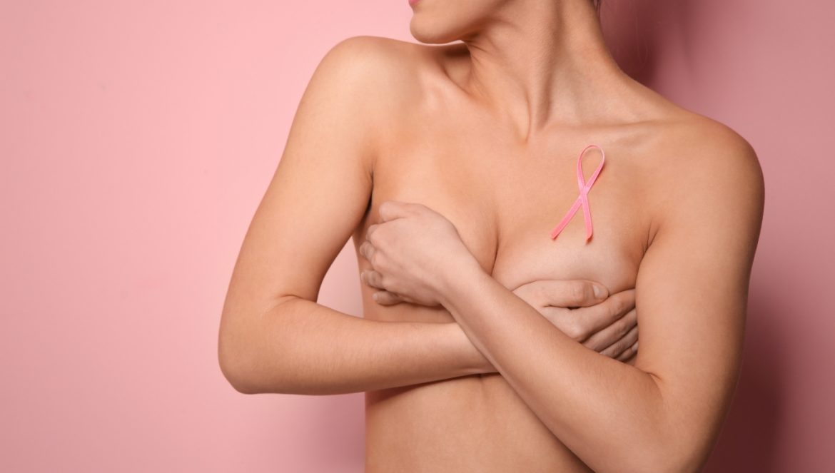 Profilaktyka raka piersi - jakie badania wykonać, aby upewnić się, że piersi są zdrowe? Kobieta bez biustonosza z różową wstążką na piersi trzyma się za biust.
