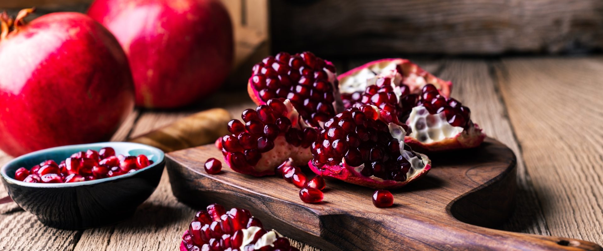 Przepisy z owocem granatu. Obrany granat na drewnianej desce do krojenia, w tle dwa całe owoce.