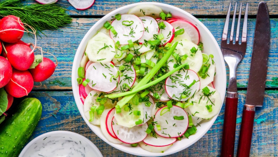 Przepisy z rzodkiewką Agnieszki Zelazko, autorki książek kulinarnych. Sałatka z rzodkiewką, szczypiorkiem i ogórkiem w misce na niebieskim stole.