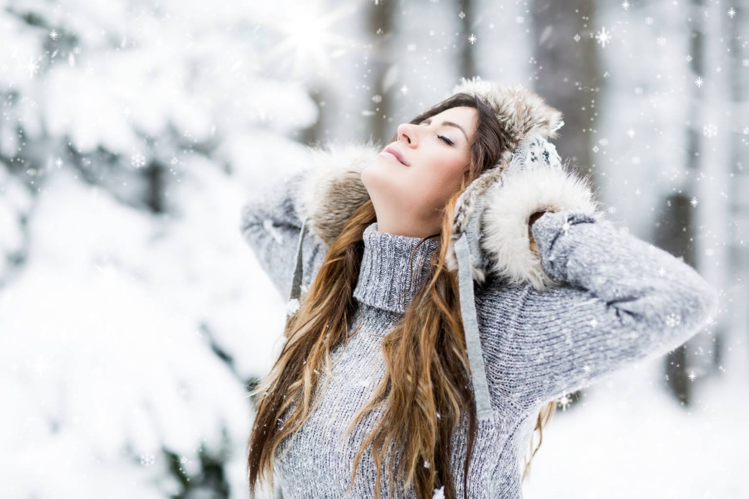 Domowe sposoby na przeziębienie. Młoda kobieta stoi na śniegu, w lesie, trzyma rękami czapkę i zadziera głowę w górę.
