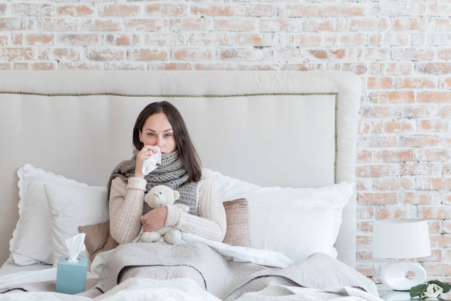 Jak poradzić sobie z uporczywym katarem? Jakie są sposoby na katar i jak szybko wyleczyć przeziębienie? Kobieta leży w łóżku w szaliku, swetrze, wyciera nos chusteczką i przytul misia.