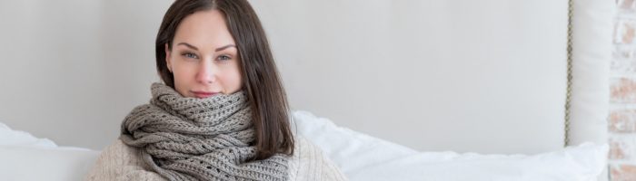 Zapalenie migdałków - objawy i leczenie. Chora kobieta z szalikiem oplecionym wokół szyi leży pod kołdrą w łóżku w sypialni.
