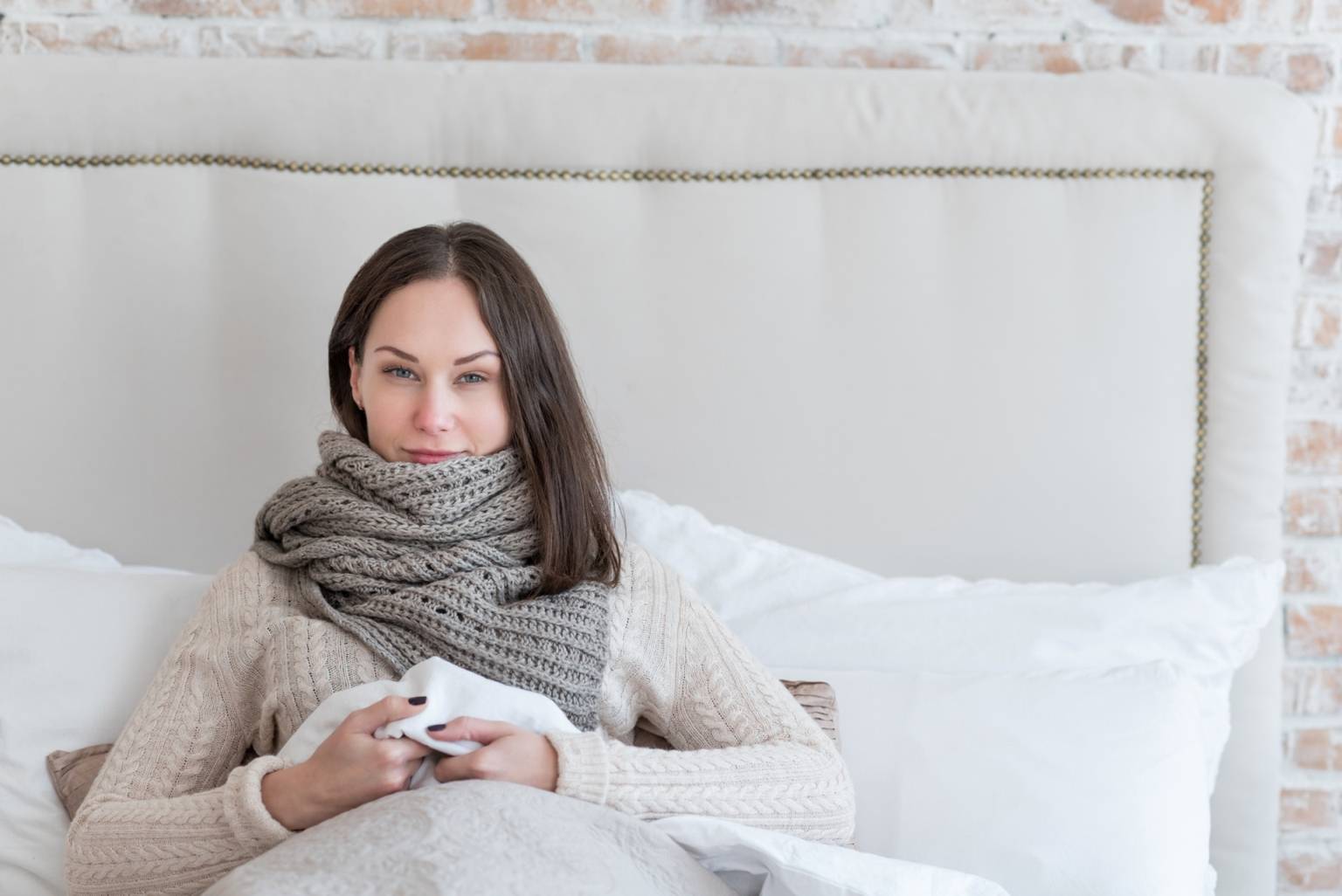 Domowe sposoby na katar i kichanie. Przeziębiona kobieta leży w łóżku pod kołdrą, opatulona szalikiem, patrze prosto w aparat i delikatnie się uśmiecha.