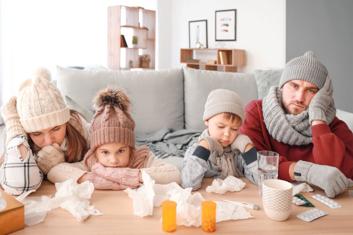 Wirusy - jak nie zachorować? Chora rodzina - mama, tata, córeczka, synek - w czapkach, szalikach i swetrach siedzą w salonie i opierają się ze zmęczenia o ławę stojącą przy kanapie.