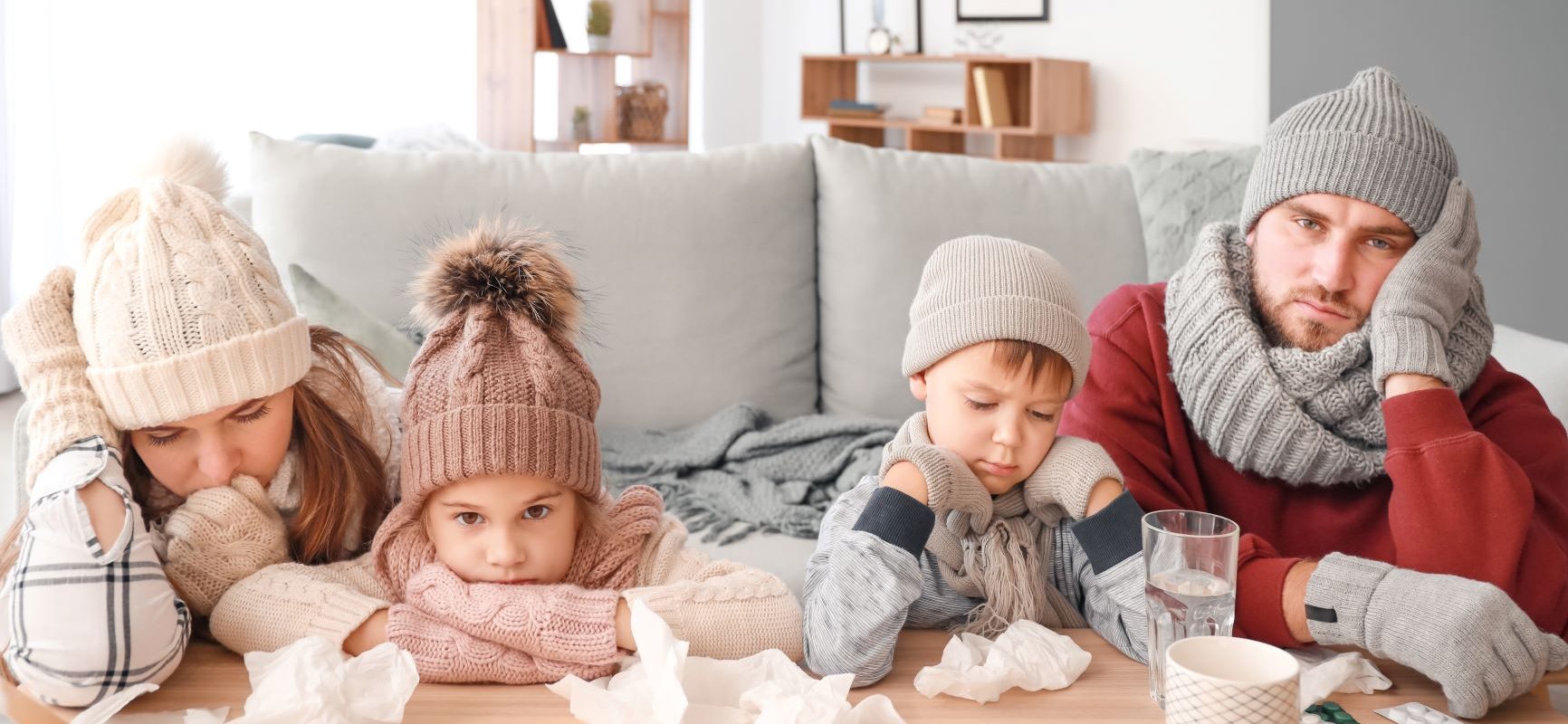 Wirusy - jak nie zachorować? Chora rodzina - mama, tata, córeczka, synek - w czapkach, szalikach i swetrach siedzą w salonie i opierają się ze zmęczenia o ławę stojącą przy kanapie.