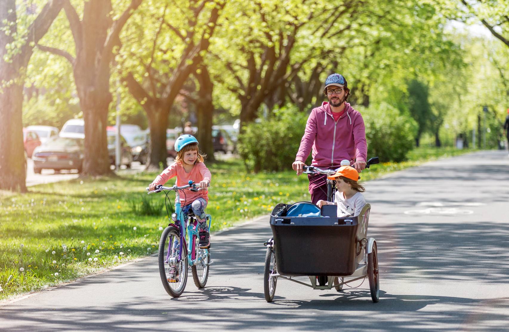 Przyczepka do roweru dla dziecka. Tata wiezie dziecko w przyczepce, obok jedzie córeczka na rowerze.