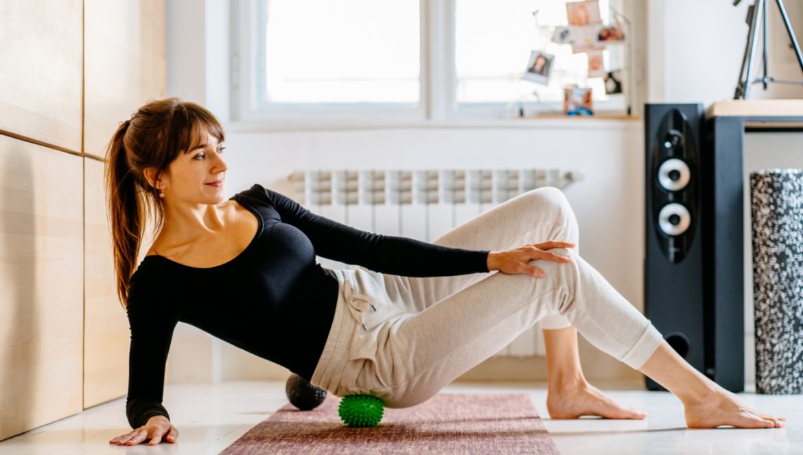 Punkty spustowe - co to jest, jak się objawia i jak sobie z tym poradzić? Kobieta rozmasowuje punkty spustowe za pomocą piłeczki do masażu, leżąc na macie w salonie w domu.