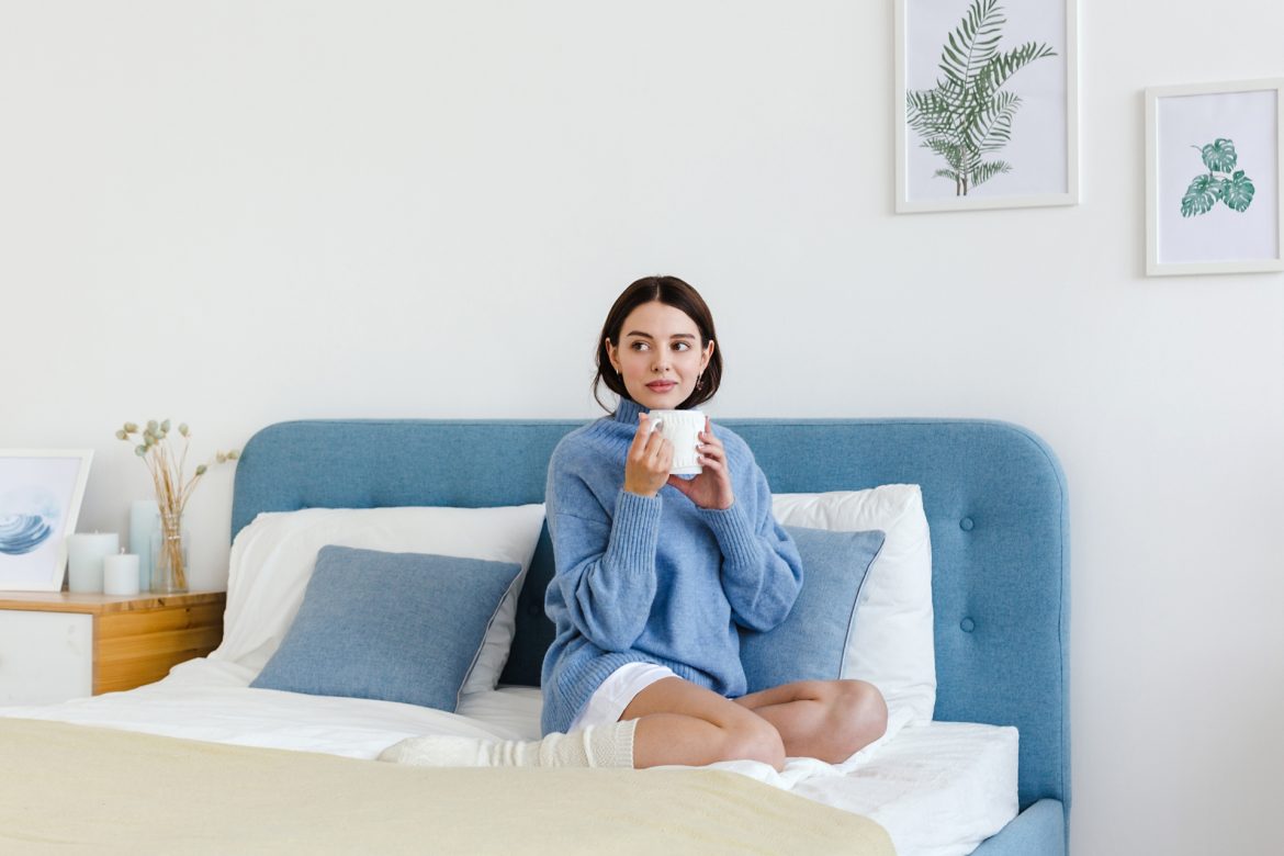 Jakie czynniki obniżają odporność? Kobieta w niebieskim swetrze siedzi na łóżku i pije gorącą herbatę.