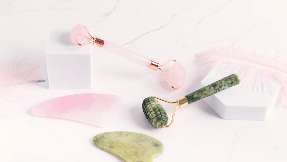 Zimno dla urody: masaż lodem, roller jadeitowy, kamień gua sha, kosmetyki z lodówki, kriolipoliza. Modne akcesoria do twarzy: roller jadeitowy i kwarcowy oraz kamienie gua sha leżą na białym blacie.