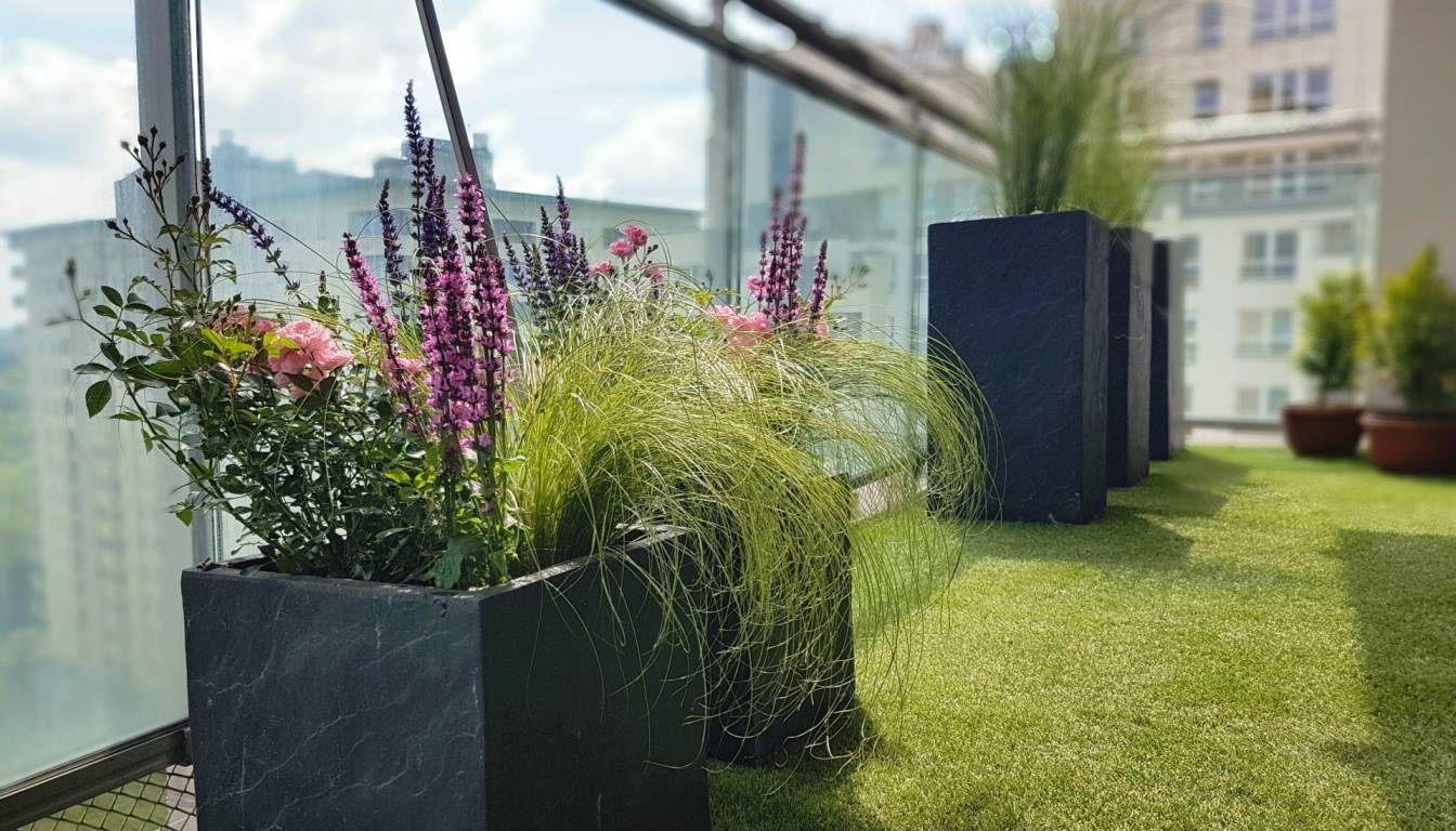 Ogródek na balkonie - jak go zaprojektować? Sztuczna trawa na balkonie i czarne donice z kwiatami.