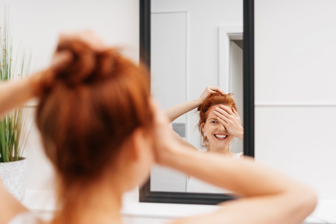 Samobadanie piersi - jak badać pierś? Rudowłosa dziewczyna uśmiecha się wstydliwie do siebie w lustrze.