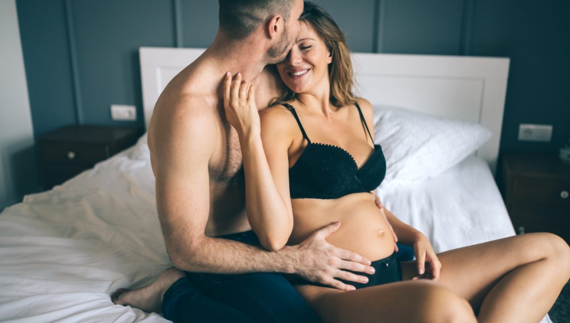 Seks w ciąży - czy można go bezpiecznie uprawiać? Zakochana para siedzi na łóżku, mężczyzn obejmuje od tyłu kobietę w ciąży w czarnej bieliźnie.