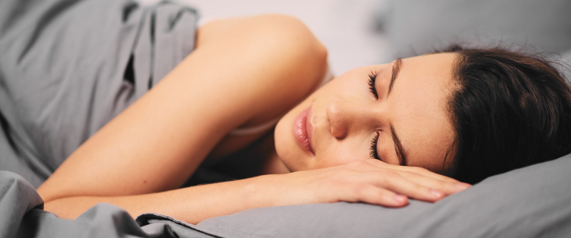 Fazy snu - jak wpływają na jego jakość? Jak zadbać o dobry sen? Kobieta śpiąca w łóżku w szarej pościeli.