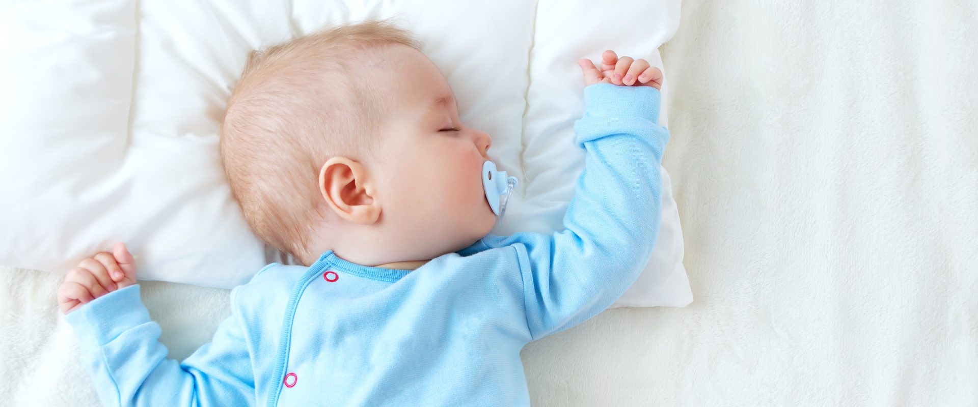 Sen niemowlaka - ile śpi niemowlę? Niemowlak w błękitnym body śpi ze smoczkiem w buzi w białej pościeli.