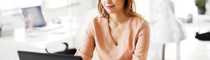 Hemoroidy - jak się pozbyć żylaków odbytu na dobre? Kobieta w łososiowej bluzce siedzi przy biurku przy laptopie.