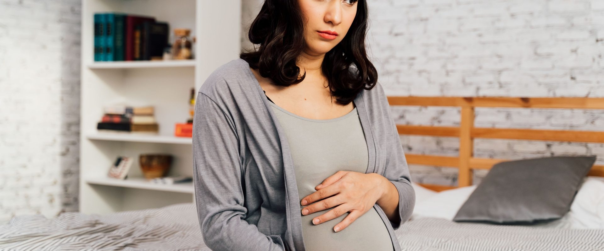 Skurcze porodowe czy skurcze przepowiadające? Jak je odróżnić? Kobieta w ciąży siedzi na łóżku i dotyka swojego brzucha.