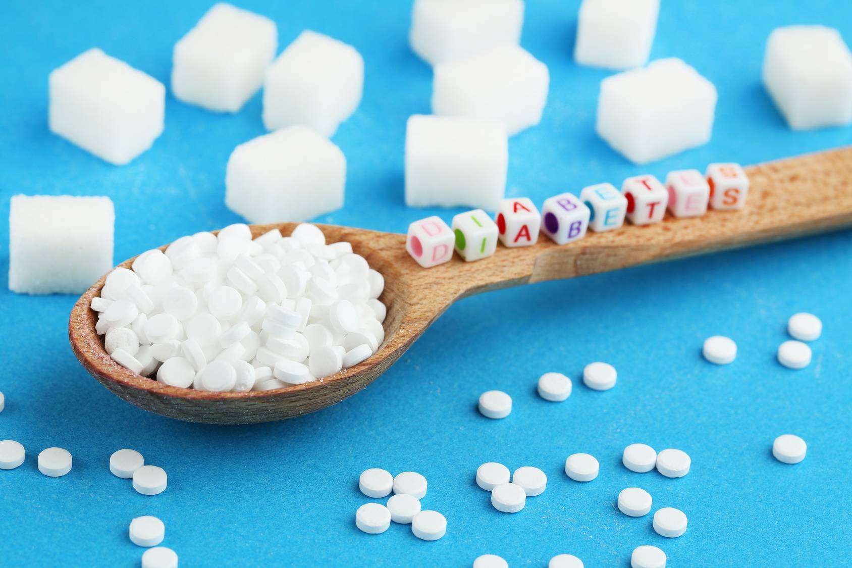 Objawy cukrzycy typu 2. Słodziki i kostki cukru na drewnianej łyżce na niebieskim tle. Na łyżce ułożony napis z klocków Diabetes.