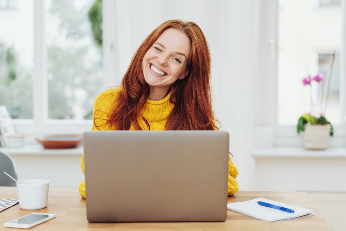 Słowa mają moc - jak wpłynąć językiem na pozytywne myślenie? Rudowłosa dziewczyna w żółtym swetrze siedzi przed laptopem i puszcza oko do aparatu.