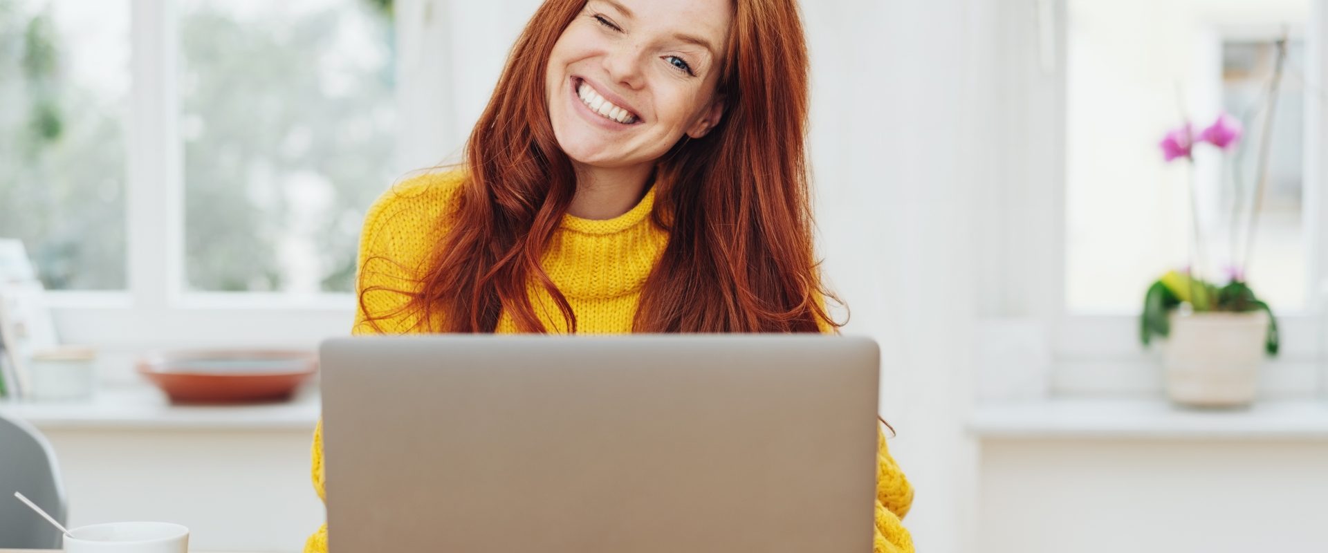 Słowa mają moc - jak wpłynąć językiem na pozytywne myślenie? Rudowłosa dziewczyna w żółtym swetrze siedzi przed laptopem i puszcza oko do aparatu.