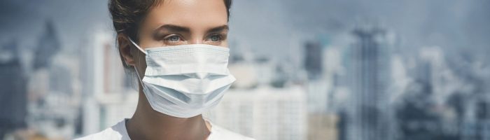 Jakość powietrza w Polsce - raport Polskiego Alarmu Smogowego 2021. Dziewczyna w białej koszulce i białej maseczce ochronnej z włosami upiętymi w kok stoi na tle wielkiej metropolii zanieczyszczonej przez smog i dymy z kominów.