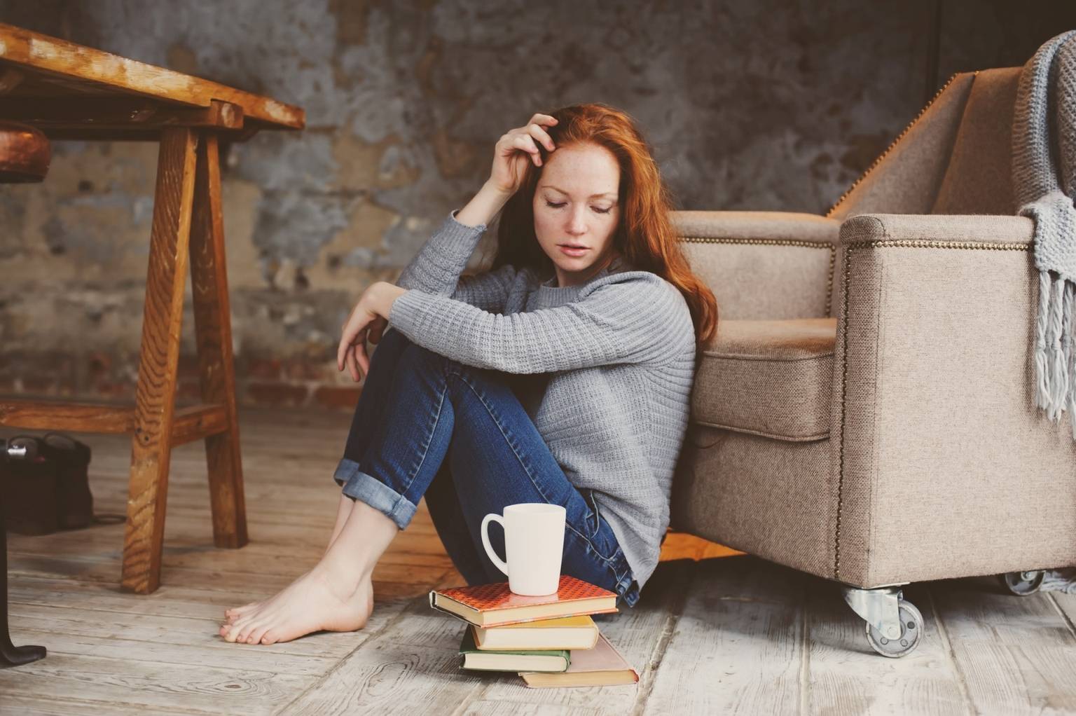 Jak leczyć depresję wg medycyny chińskiej? Rudowłosa zamyślona dziewczyna w szarym swetrze i dżinsach siedzi w pokoju na podłodze, opiera się plecami o fotel, patrzy na kubek herbaty i książki.