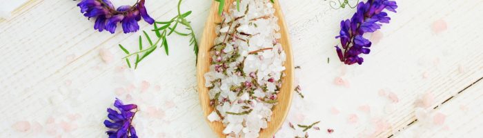 Sól gorzka, sól Epsom do kąpieli i roztworów. 5 sposobów na wykorzystanie soli Epsom. Gruboziarnista sól na drewnianej łyżce, obok leżą liście i fioletowe kwiaty.