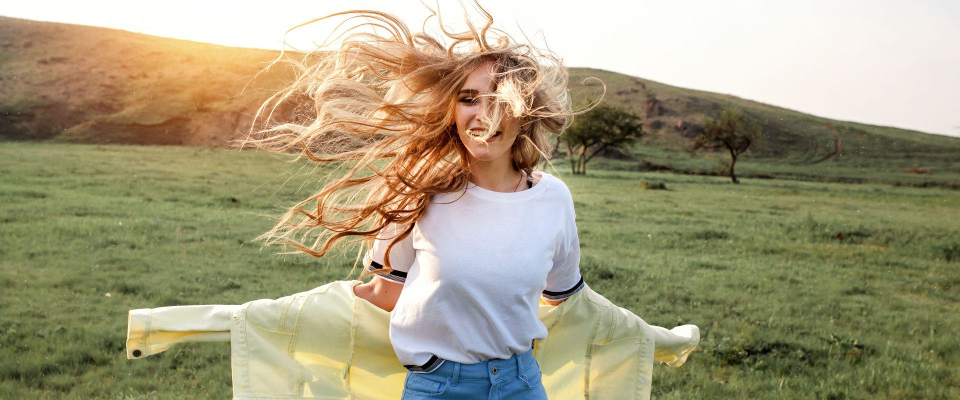 Wiosenne SPA dla włosów, czyli jak pielęgnować włosy wiosną - Kobieta na zielonym wzgórzu z rozwianymi na wietrze blond włosami uśmiecha się do aparatu.