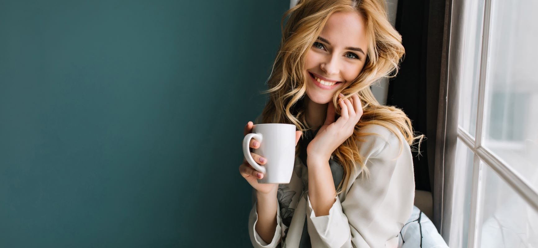 Sposoby na chandrę, czyli skąd czerpać energię do życia. Uśmiechnięta kobieta w blond włosach i piżamie pije poranną kawę, siedząc na parapecie.