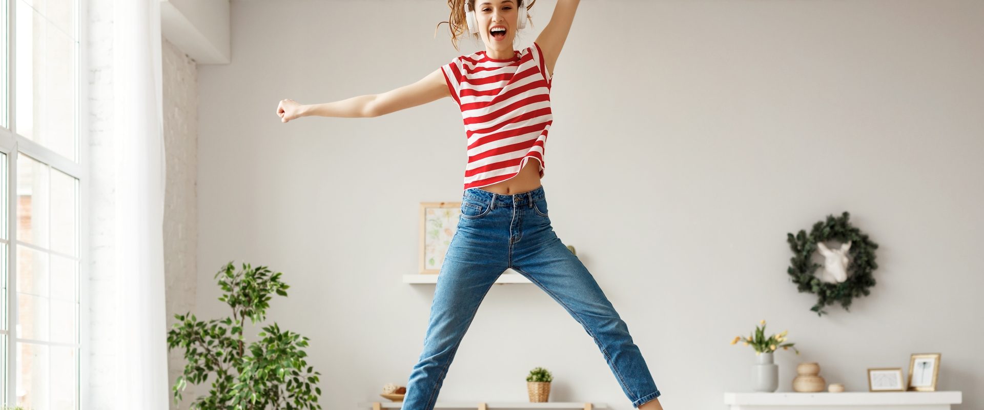 Sposoby na dobry nastrój. Młoda dziewczyna w bluzce w czerwono-białe paski, dżinsach i czerwonych trampkach i słuchawkach na uszach, skacze na kanapie w salonie.