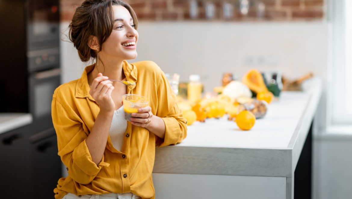 10 sposobów na utrzymanie prawidłowego poziomu glutationu w organizmie. Młoda kobieta z żółtej bluzce je pudding z chia, opierając się o blat kuchenny.