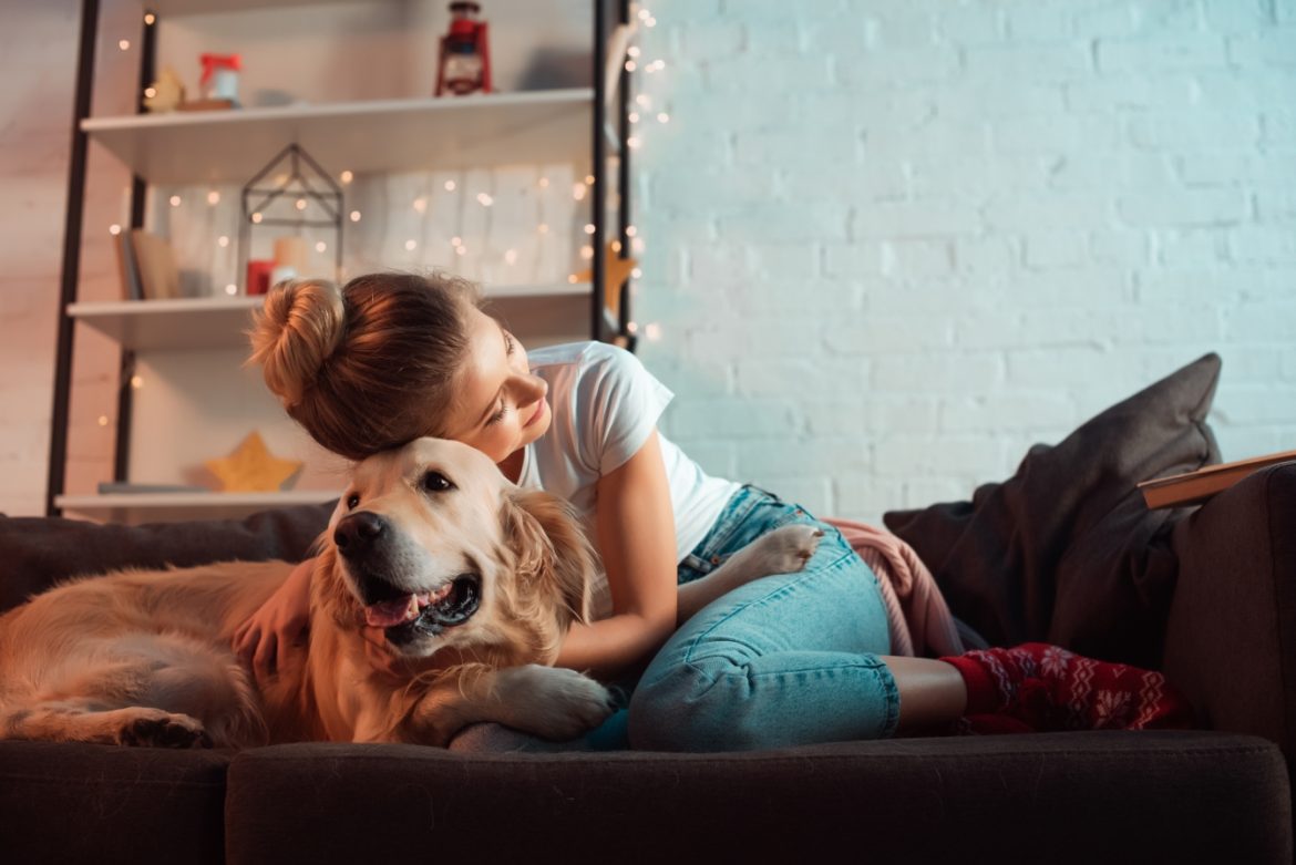 Miłość singielki - dobre życie bez partnera. Wywiad z psycholog Joanną Kot. Młoda kobieta w dżinsach i koszulce przytula się do swojego psa golden retrivera, siedząc na kanapie w domu.