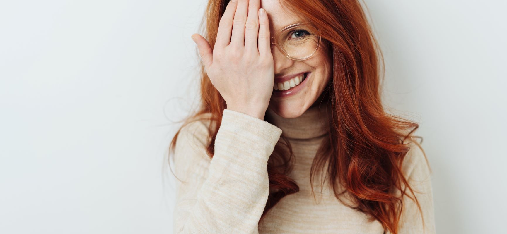 Domowe sposoby na zmęczone oczy. Młoda kobieta o rudych włosach w okularach przykłada dłoń do jednego oka i się uśmiecha do aparatu.