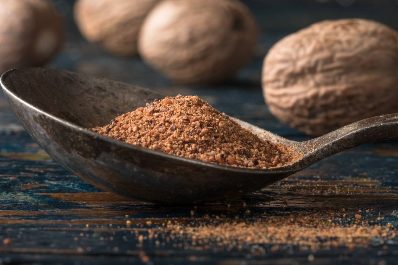 Gałka muszkatołowa mielona na łyżeczce, w tle leżą orzechy muszkatołowca.