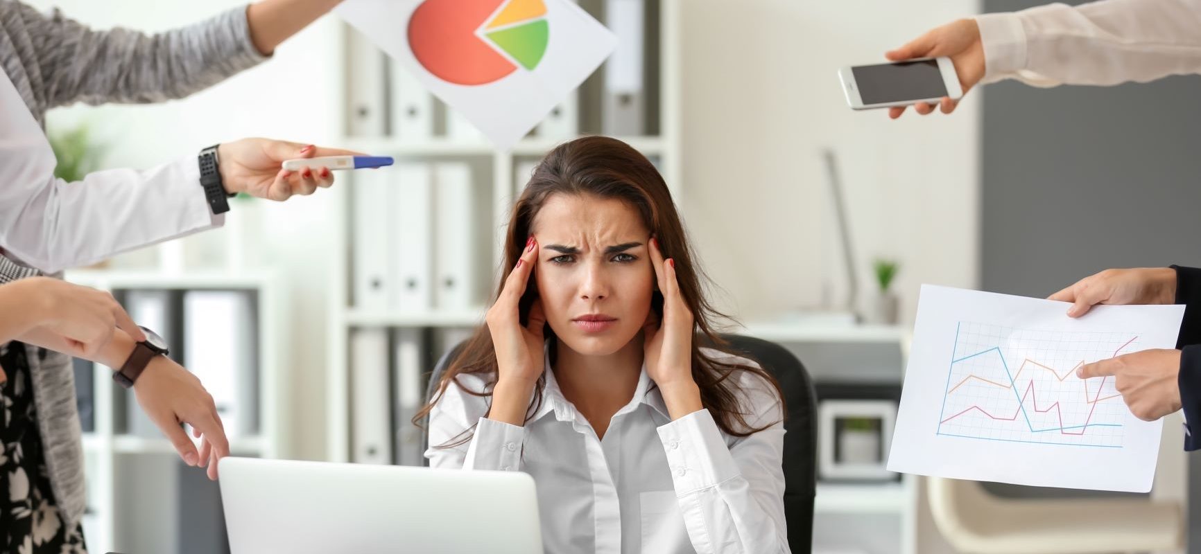 Stres w pracy - jak sobie z nim poradzić? Kobieta w białej koszuli siedzi w przy biurku w pracy i trzyma się za głowę przytłoczona obowiązkami, ktoś z każdej strony czegoś od niej chce.