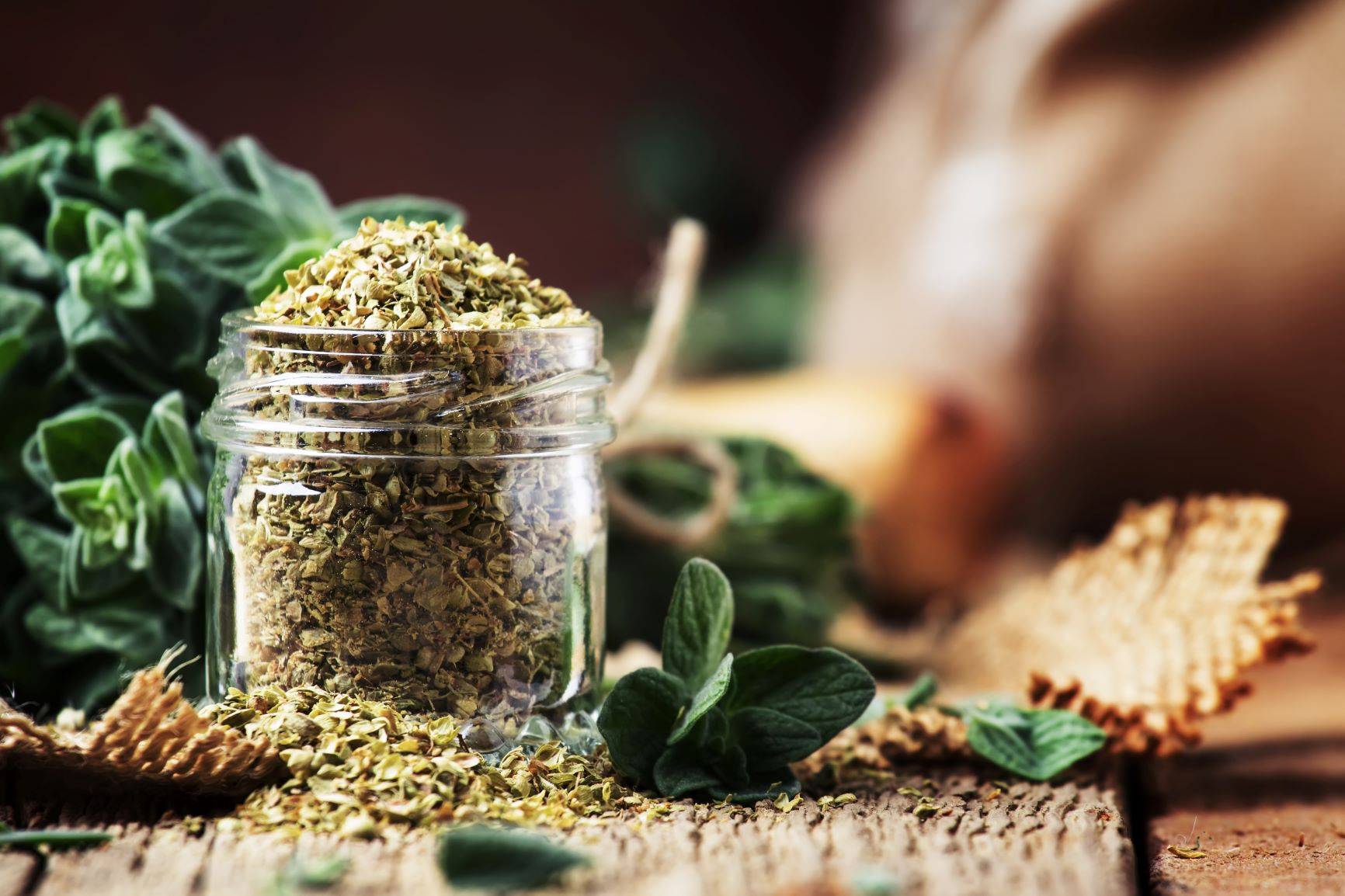Herbatka z oregano pomaga zwalczać wirusy, bakterie i pasożyty. Suszone oregano w słoiczku na drewnianym stole, obok leżą listki lebiodki.