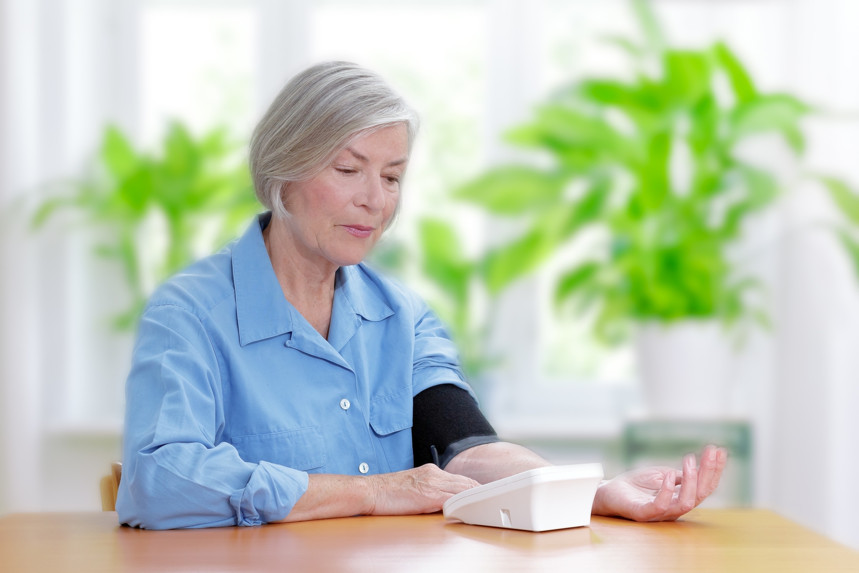 Światowy Dzień Nadciśnienia Tętniczego. Jak prawidłowo mierzyć sobie ciśnienie krwi? Seniorka mierzy ciśnienie ciśnieniomierzem w domu, opierając rękę na stole w salonie.