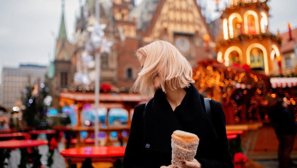 Kulinarna podróż po wigilijnej Polsce - jakie potrawy wigilijne jadamy w różnych regionach kraju? Młoda kobieta w krótkich blond włosach stoi na rynku we Wrocławiu ze świątecznym ciastkiem z jarmarku w ręku.