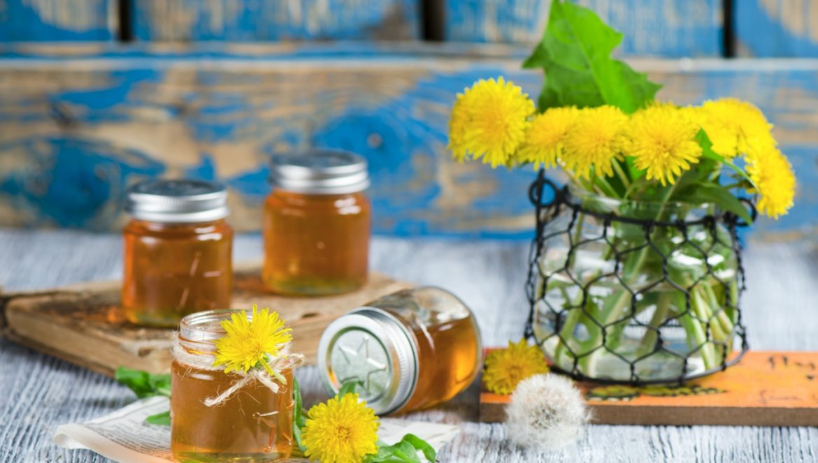 Syrop z mniszka lekarskiego - sprawdź przepis! Małe słoiczki z syropem z mniszka na rustykalnym niebieskim drewnianym blacie, w tle stoi wazon z kwiatami mniszka.